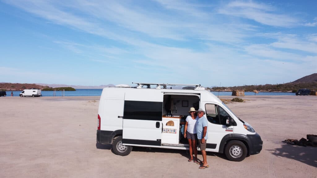 A man and woman pose outside of the camper van parked on a beach